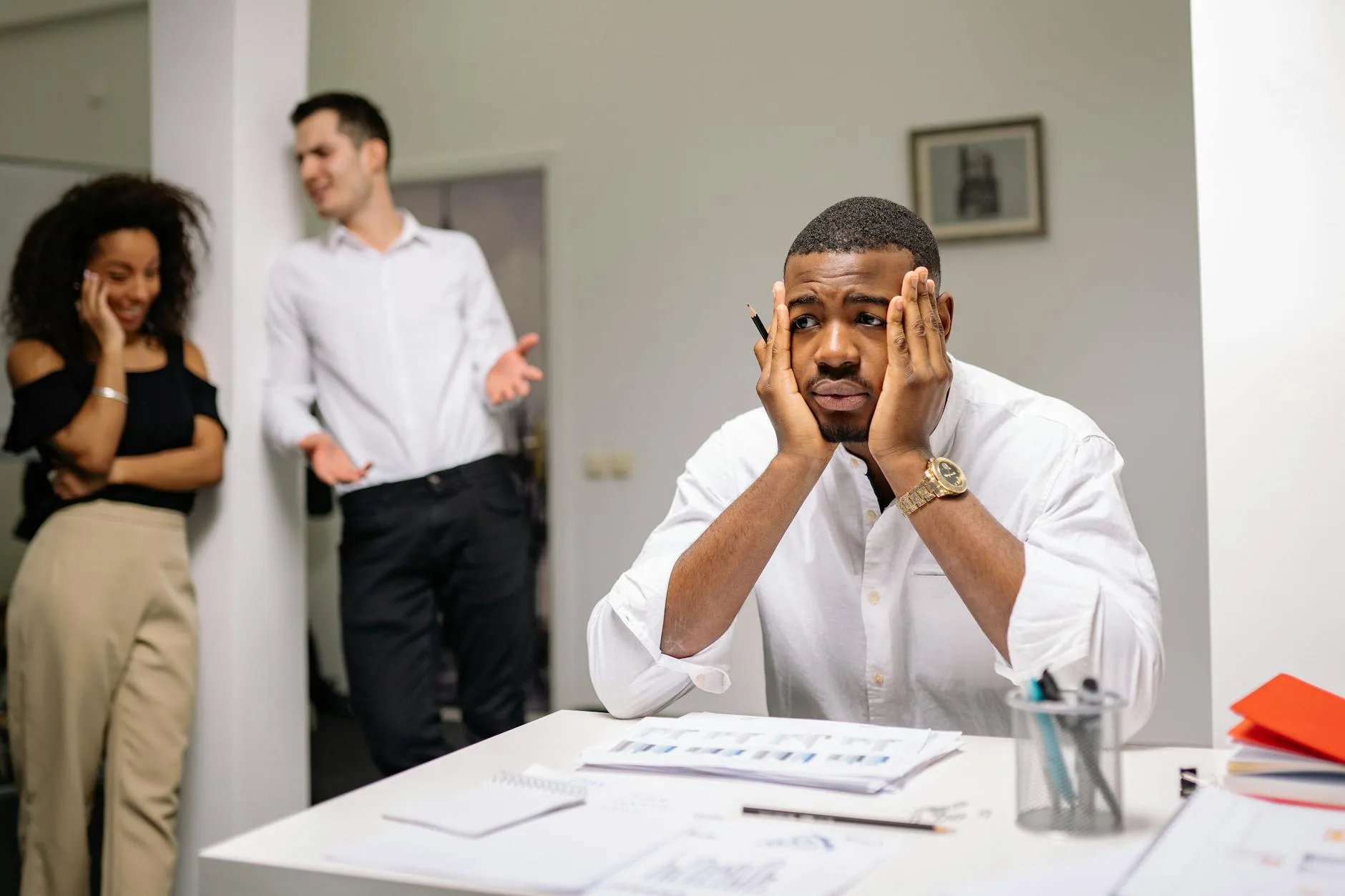 A stressed employee sits at a desk with colleagues in discussion nearby, highlighting workplace tension.
